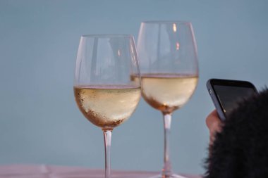 Masada beyaz şarap ve elinde cep telefonu olan siyah elbiseli bir kızın eli olan iki bardak..