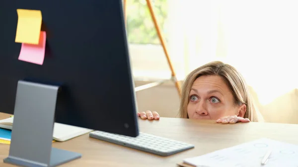 Portrait of surprised woman peeping out table and looking at pc screen. Office worker hiding under desk