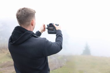 Sisli çayırda fotoğraf çeken genç bir erkek fotoğrafçı. Adam kameradan bakar, boşluğu metin için kopyalar.