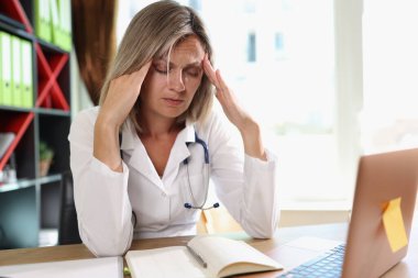 Exhausted female doctor having headache while working at clinic. Tired and depressed sad doctor with closed eyes at medical office desk.