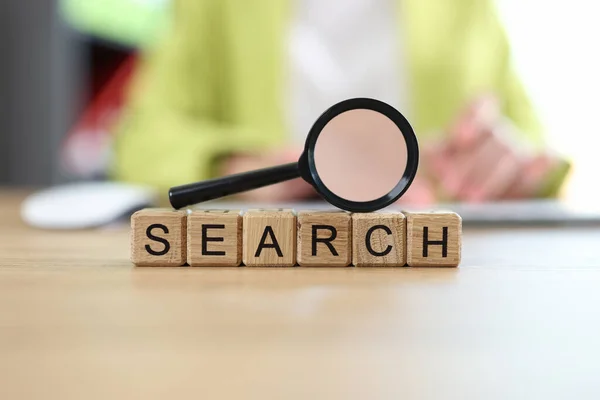 Word search on wooden cubes with magnifying glass on table, blurred person in background. Searching information, vacancy and solutions concept.