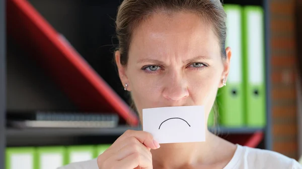 Unhappy woman in office holds paper with sad emotion in front of her mouth. Business failure and dismissal concept.