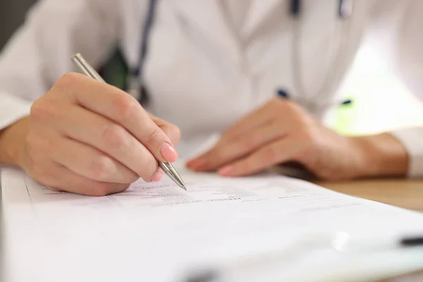 Female doctor fills out medical forms while sitting at her desk in clinic. Therapist hands with pen on medical papers close up.