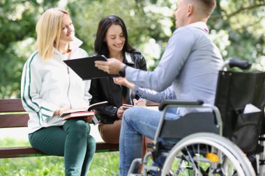 Two female students and one male student study together in park, man is sitting in wheelchair. Friends with books and tablet have fun and talking together outdoors.