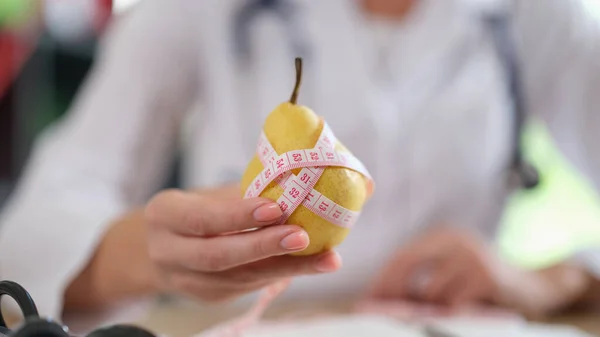 Doctor suggest eating fruits and food with vitamins. Nutritionist showing ripe yellow pear with measuring tape while sitting in medical office.