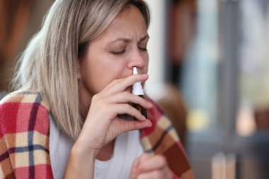 Woman with illness, covered in blanket, uses nasal spray close-up. Cold season and weak immunity concept.