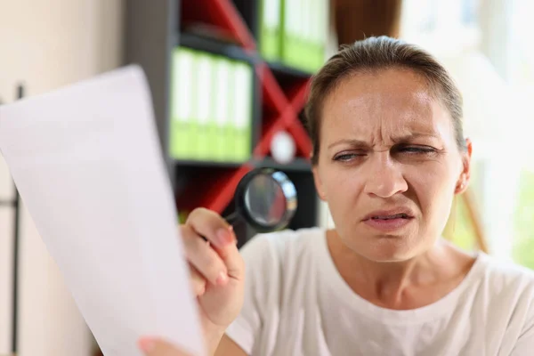 Incredulous middle age woman looking on document trough magnifying glass close-up. Concept of eye diseases and people incredulity.