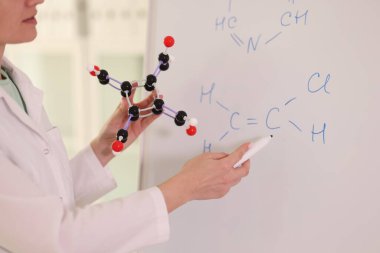 Female chemist holding molecular model and drawing chemical formulas on whiteboard. Chemistry, education and science research concept.