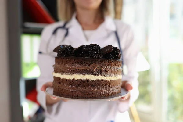 Female doctor advises stop eating high calorie unhealthy sugary foods. Dietologist holds whole chocolate cake and warns against overeating.