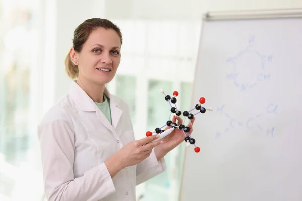 Female scientist standing near whiteboard, pointing to molecular model in her hand and looking at camera. Scientific research and education concept.