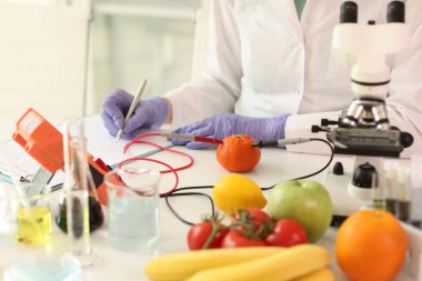 Scientist with microscope and research equipment takes notes while examining many fruits and vegetables. Concept of checking quality and properties of food samples.