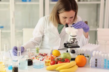 Female scientist with microscope, test tubes, laboratory glassware and reagents examining many fruits and vegetables in her lab. Dieting and food quality research concept.