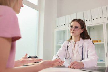 Young doctor listens to her patient while sitting at medical clinic table. Female patient talks about symptoms of diseases to doctor in hospital.