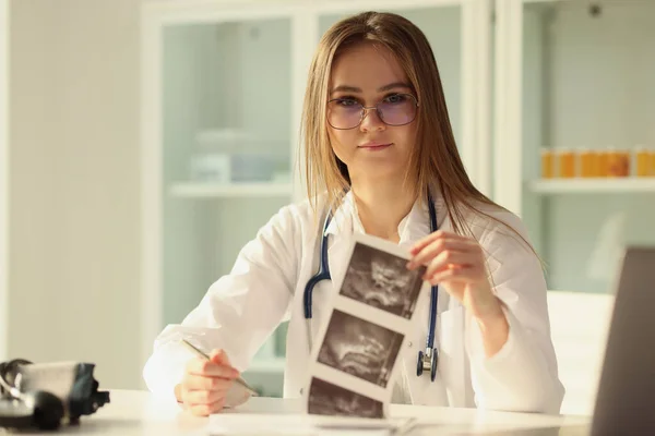 Young female doctor working with ultrasound scan results and looking at camera. Nurse working with medical documents while sitting at desk in medical clinic.
