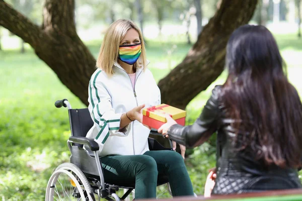 Woman with illness in wheelchair receives box with gift from her friend in park. Caring for disabled, supporting friends and togetherness concept.