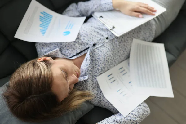 A young woman sleeps in office on couch covered with documents, close-up, above. Fatigue, deadline