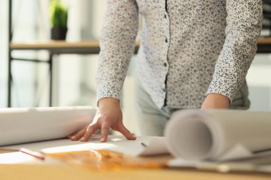 Female designer is standing near her work desk with rolls of paper. Architect works in her office with projects close-up.