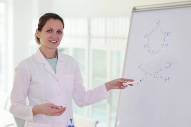 Gülümseyen kadın kimyager beyaz tahtadaki kimyasal formüllere işaret ediyor. Eğitim ve bilimsel araştırma kavramı.