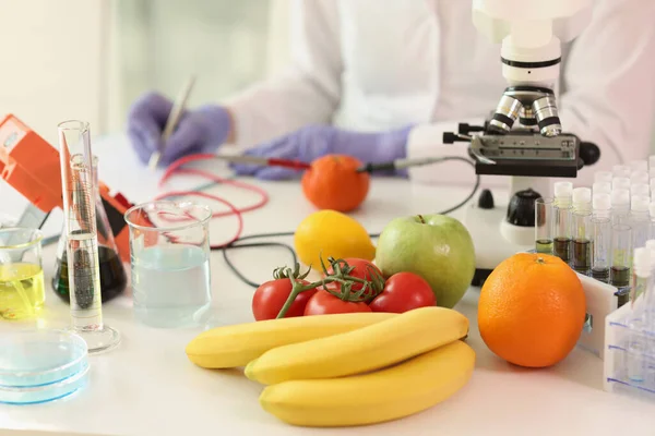 Fruits and vegetables with scientific equipment on table of research laboratory, scientist takes notes in background. Concept of food quality and plant properties.