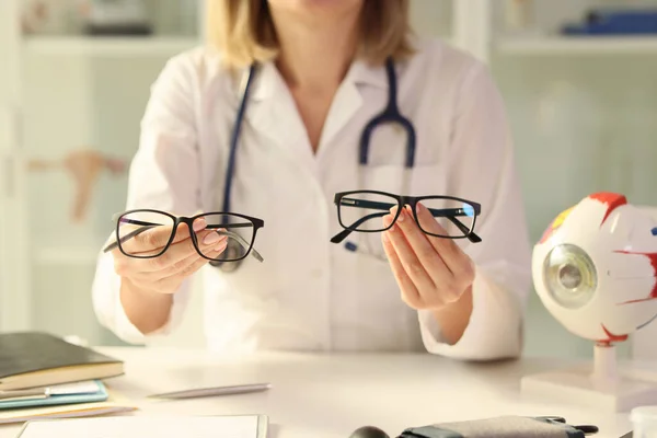眼科医は診療所の机に座っている間 さまざまな眼鏡を手に持っています 医者は患者にメガネを推薦する — ストック写真