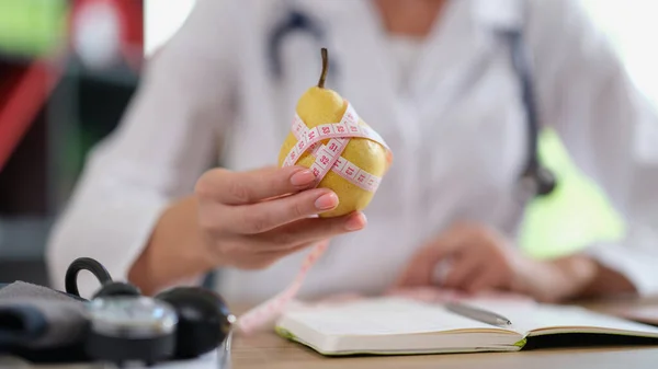 Dietologist suggest eating fresh fruits as source of vitamins. Female nutritionist holds pear and measuring tape while sitting in medical clinic.