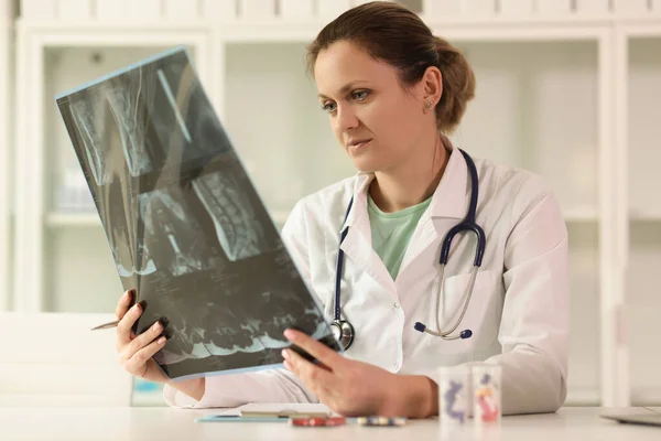 聴診器を持つ医師は オフィスのテーブルに座っている患者の背骨のX線を見て分析します 女性は診療所で放射線画像を調べる — ストック写真