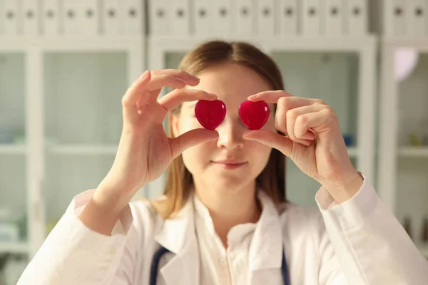Young female doctor holds red hearts with hands against eyes. Positive woman in professional uniform in clinic office. Heart health care