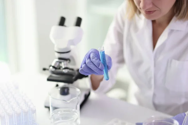 女性労働者は顕微鏡を用いて試験管内のサンプルを調べる ゴム手袋の女性は研究室の密室で科学研究を行うテーブルに立っている — ストック写真