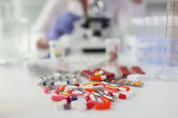 顕微鏡で研究を行う女性に対して白いテーブルの上に散在する錠剤の山 研究室での医学 科学研究 — ストック写真