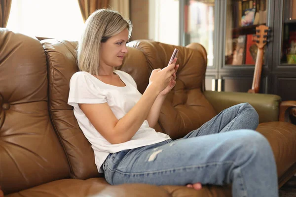 女性は快適なソファに座ってスマートフォンを介してソーシャルネットワーク上の写真を投稿します ブロンドの女性は仕事の後家で過ごすことを楽しむ — ストック写真