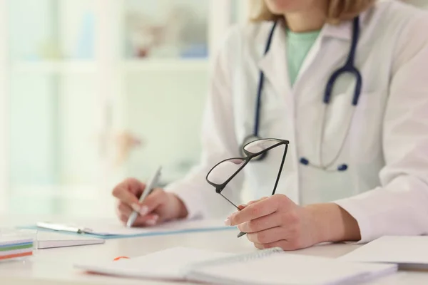 女医は病院の診療所の近くのテーブルに眼鏡をかけたメモを書いている セラピストは職場で患者の医療記録を記入します — ストック写真