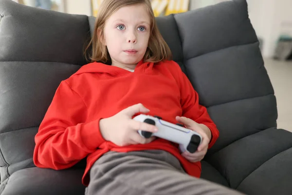 Focused teenager girl in red hoodie holds gaming console making tense facial expression. Child plays video games after school at home closeup