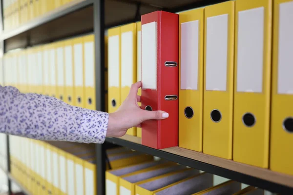 Hand of woman taking red ring binder among yellow items put in row on shelf. Organized folder structure with archive and materials in office closeup