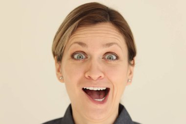 Aktif yüz ifadeleri olan bir kadın kameraya bakıyor. Kadın gözleri şişer ve bej arka planda dikilirken ağzını açar.