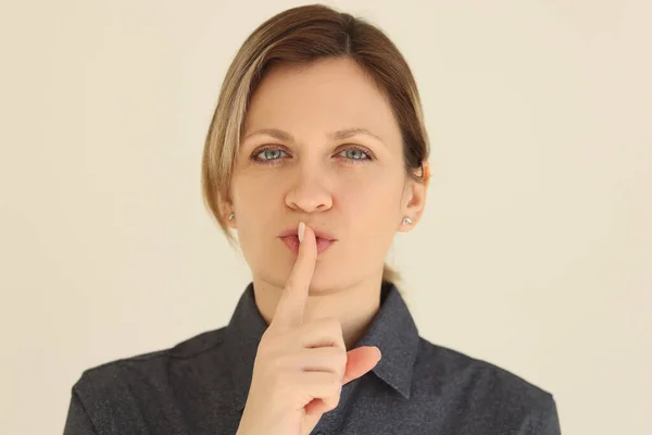 Woman puts index finger to mouth asking to keep silence. Blonde female person makes silent gesture standing on beige background closeup