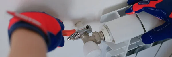 Close-up of plumber in protective gloves installing heating radiator in apartment or house. Builder installs new hot water central heating system using wrench