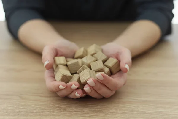 Hands of woman with neat manicure holding small wooden cubes. Female person sits at wooden table showing handful of children building toy blocks