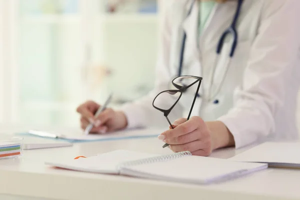 制服の女医は クリップボードにペンでノートを作る眼鏡を保持しています 聴診器を持つ女性は 治療計画を書き込みますテーブルに座って — ストック写真
