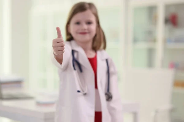 穿着军服的可爱的小女孩露出大拇指向上的姿势 带着听诊器的模糊儿童站在医院的办公室里 微笑着用相机看着 — 图库照片