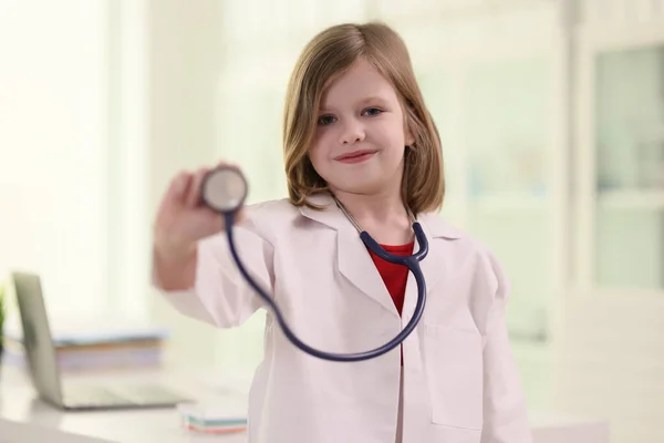 可爱的金发女孩拿着听诊器检查病人的游戏疗法 身穿医疗制服的小医生站在诊所的办公室里 看着镜头 面带微笑 — 图库照片