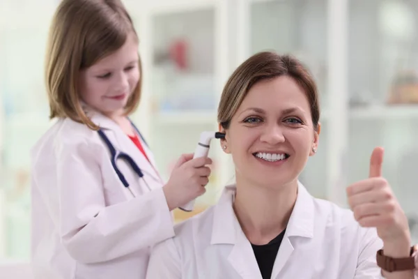 穿着医生制服的小女孩用医疗设备检查病人的耳朵 女人翘起大拇指笑着表示赞成预约时的检查 — 图库照片