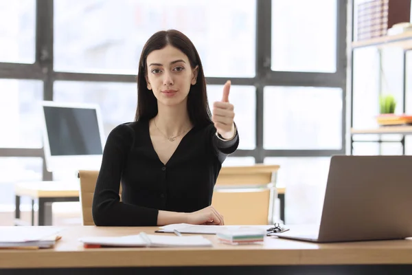 成功したマネージャーは 親指を手でジェスチャーを示す開いているラップトップの近くのテーブルに座ります ブルネットの女性は広々としたオフィス前提で仕事を承認します — ストック写真