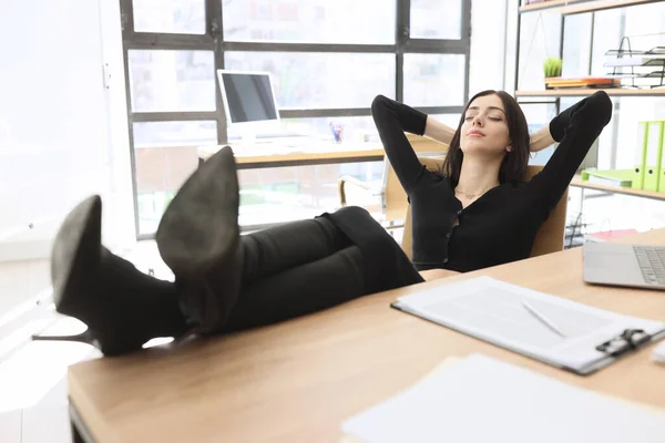 エレガントなブルネットの女性はオフィスのテーブルの上にハイヒールの靴に足を置く椅子に座っています 若いマネージャーは職場で書類作成を中断する — ストック写真
