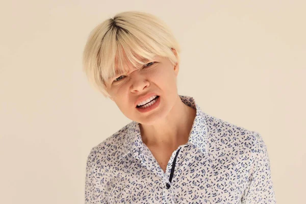 Blonde woman with short haircut opens mouth showing healthy teeth. Stylish female person in blouse squints eyes looking in camera on beige background