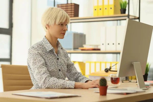 Focused woman with dyed blonde hair works on computer sitting at wooden table in office. Female manager in elegant outfit busy writing report