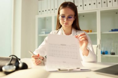 Gözlüklü genç bir kadın laboratuardaki masada oturan gazetelere bakıyor. Bilimadamı belgeleri analitik araştırma sonuçlarıyla gözden geçiriyor