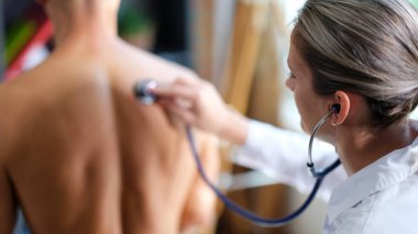 Terapist steteskop kullanarak nefes alan adamı dinliyor. Kadın doktor klinikteki hastayı arka planda bulanık bir şekilde muayene ediyor.