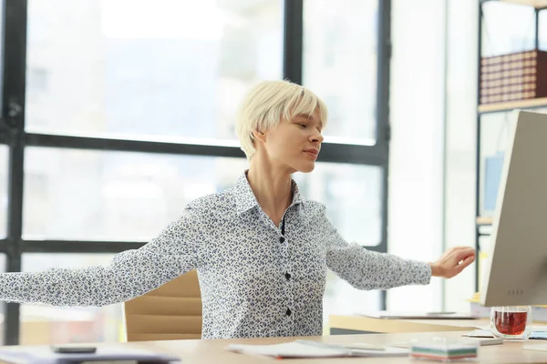 Woman stretches back and shoulders sitting at desktop in company office. Young manager warms up muscles at workplace. Healthy posture