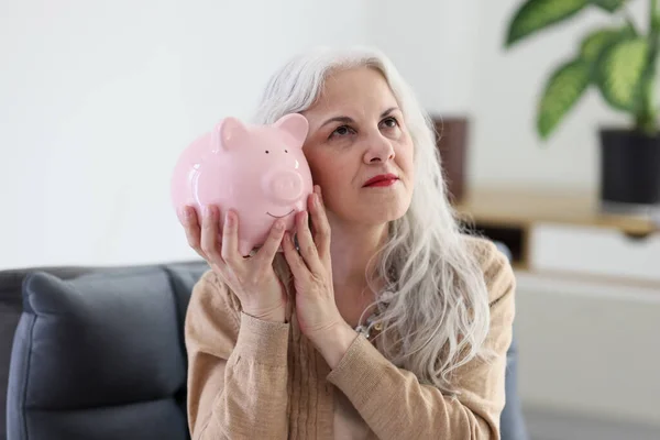 Senior Woman Put Ear Piggy Bank Sitting Armchair Home Stará — Stock fotografie