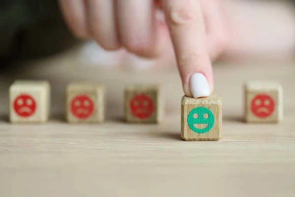 Finger of woman moves out wooden cube with green positive emoji against blocks with negative faces. Difference in moods and attitude
